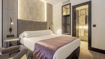 Habitación de hotel moderna con una cama grande, una mesita de noche pequeña, una silla y un baño visible a través de una puerta abierta.