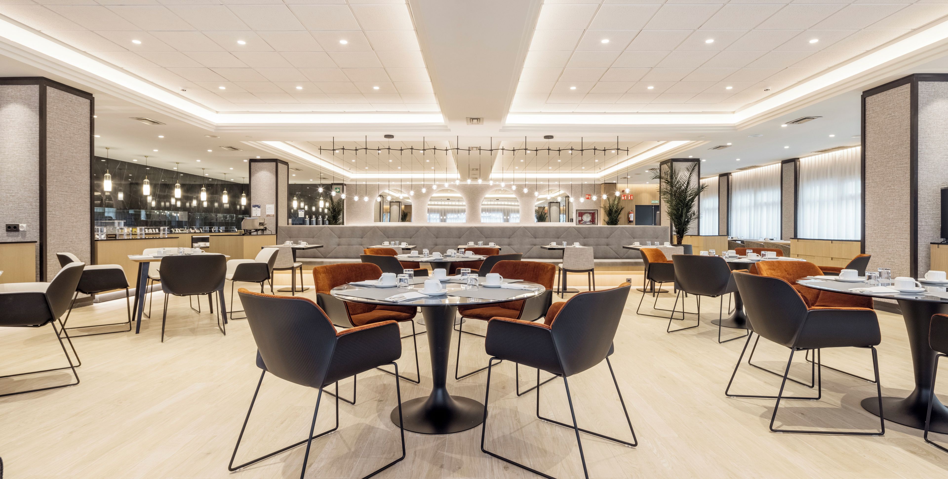 Un comedor moderno con mesas y sillas redondas, una zona de bar con mostrador, y un área de estar con techos altos y iluminación empotrada.