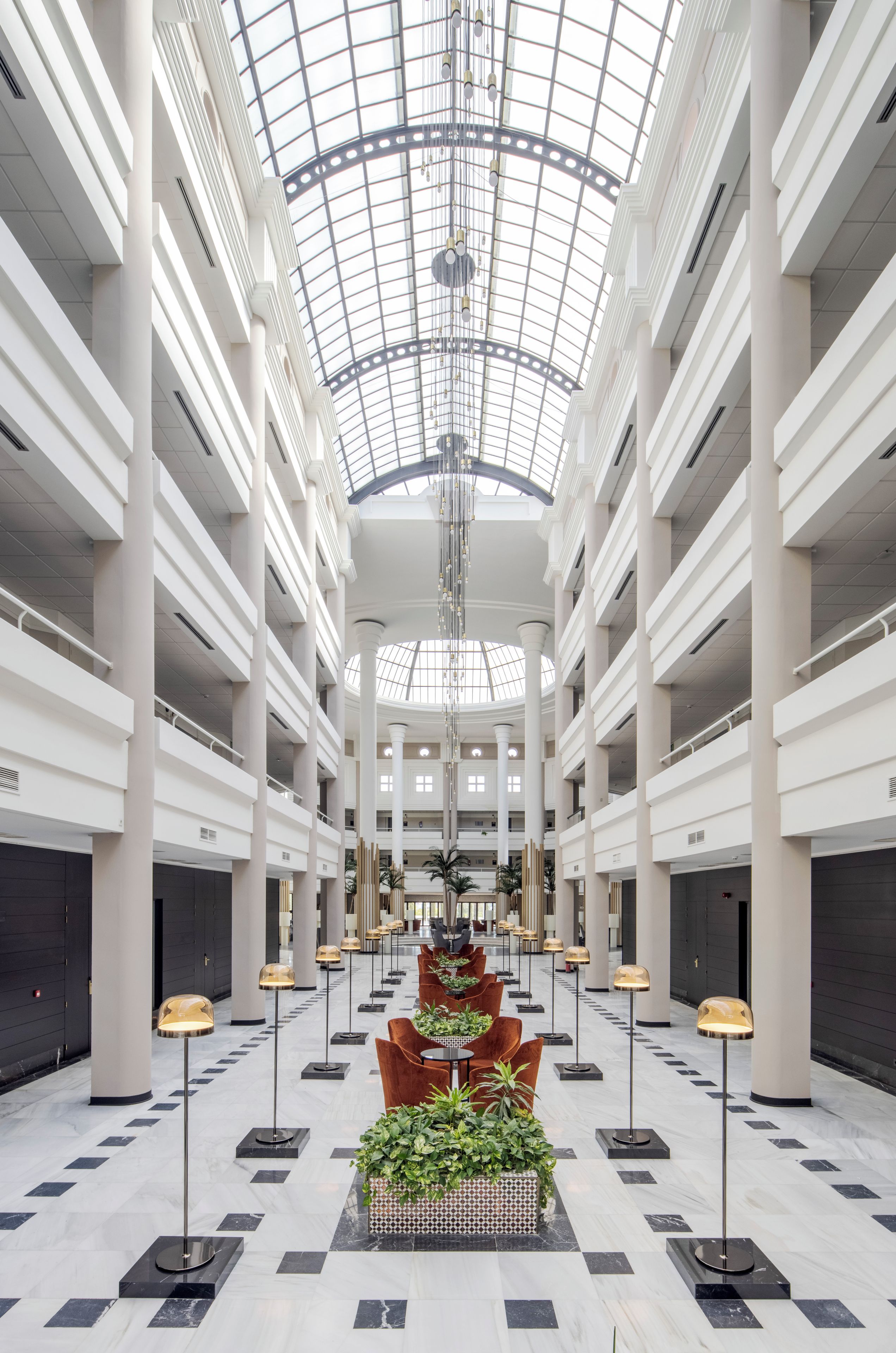 Een ruime atrium met een hoge plafond en een glazen koepel, met een centraal zitgedeelte met tafels en stoelen, omringd door zuilen en potplanten.