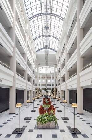Un vaste atrium doté d’un haut plafond et d’une coupole de verre, comprenant un espace de détente central avec des tables et des chaises, entouré de colonnes et de plantes en pot.