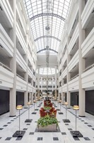 A spacious atrium with a high ceiling and a glass dome, featuring a central seating area with tables and chairs, surrounded by columns and potted plants.
