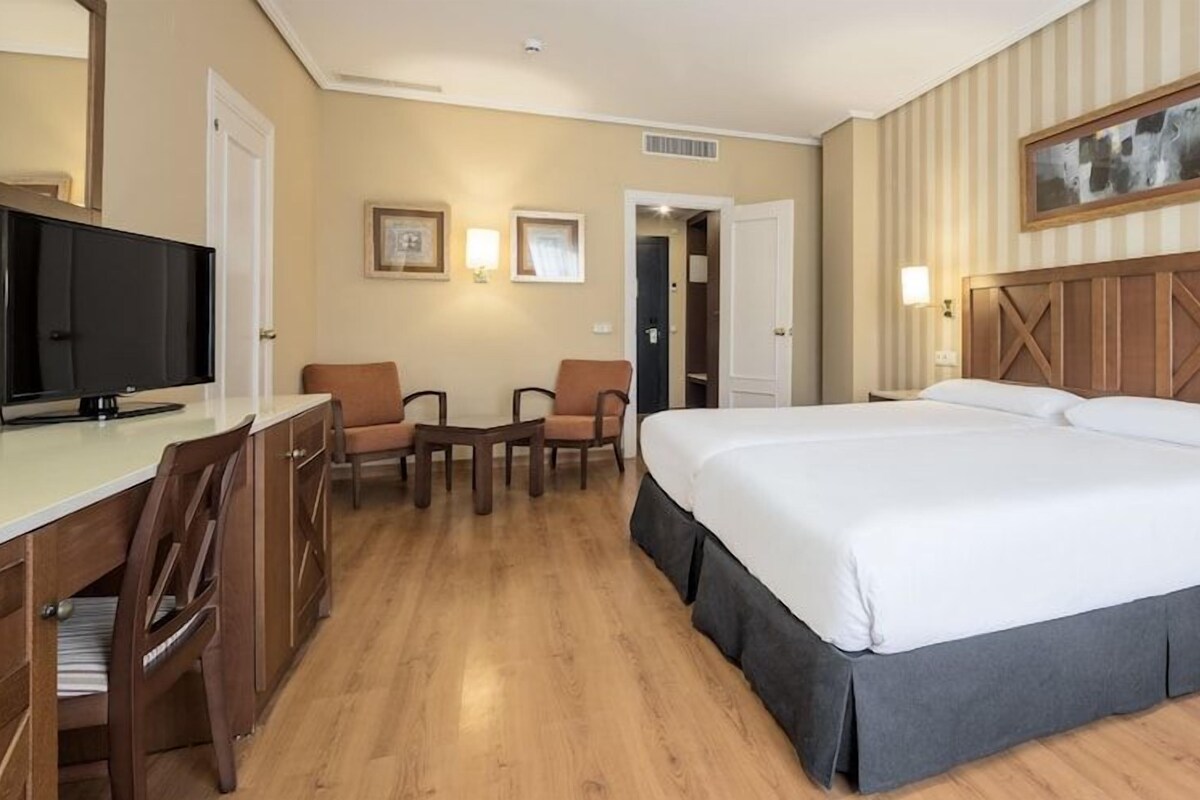 Une chambre d’hôtel avec un grand lit, un bureau avec une télévision, deux chaises et une table de chevet avec une lampe.