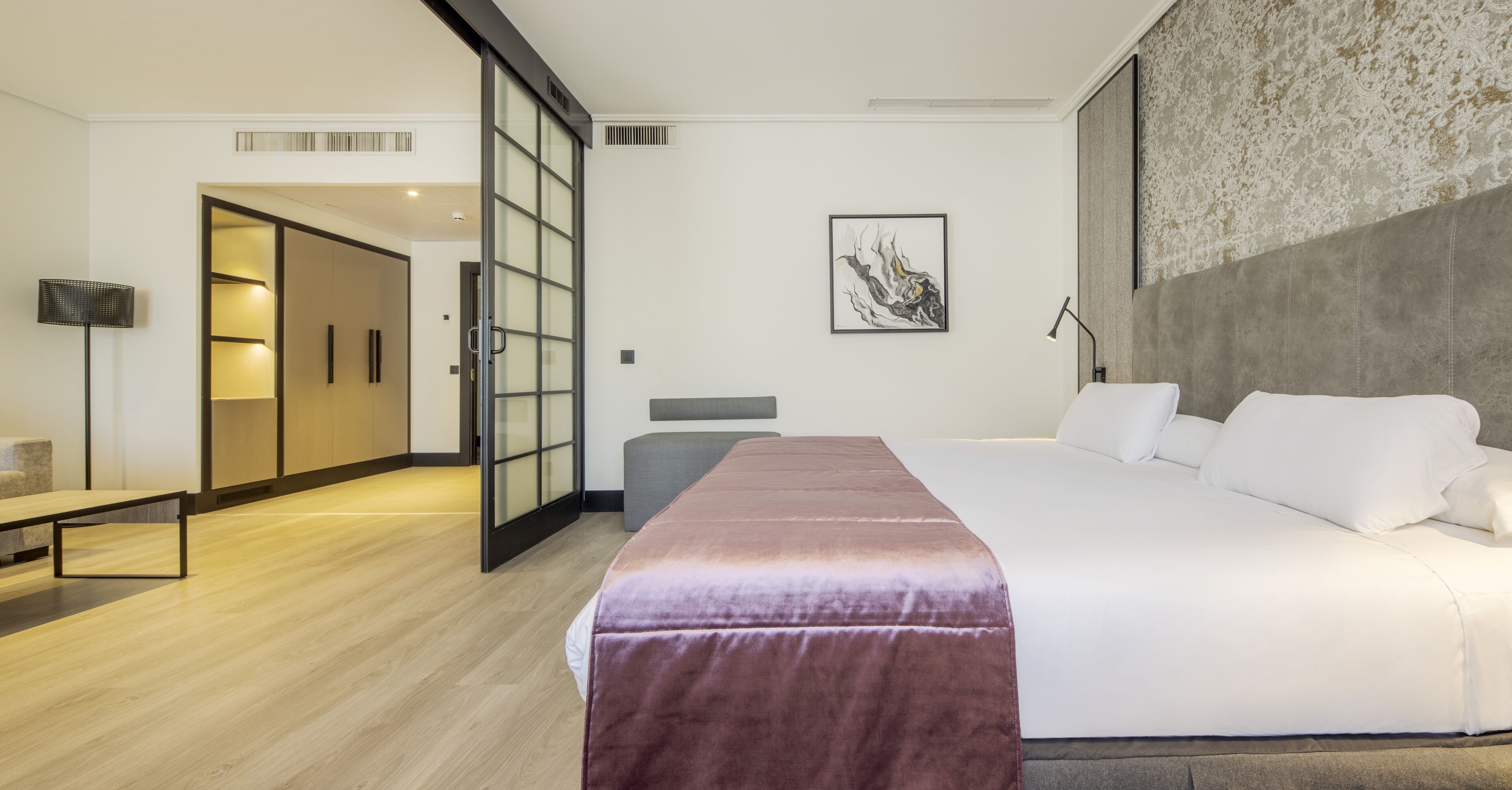 A modern hotel room with a large bed, a bench, a desk, and a sliding door leading to a bathroom.