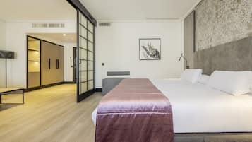 A modern hotel room with a large bed, a bench, a desk, and a sliding door leading to a bathroom.