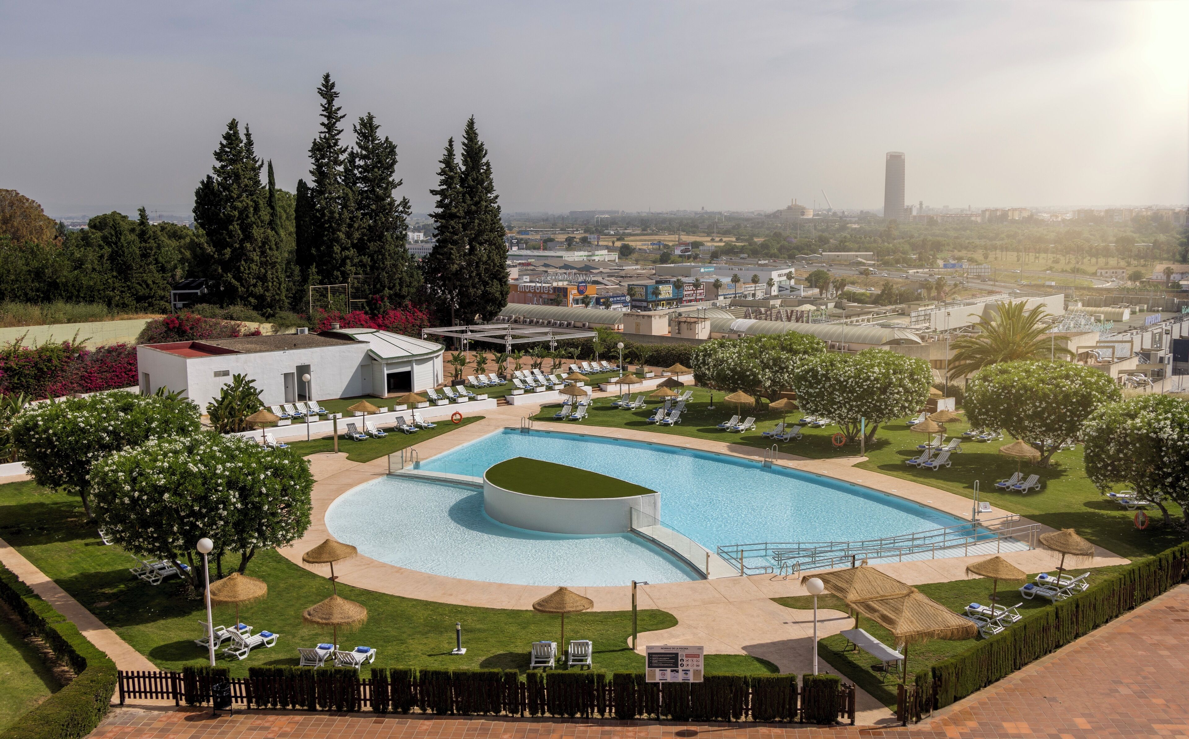 Un área de piscina con sillones de descanso, un edificio pequeño y una vista del paisaje urbano al fondo.