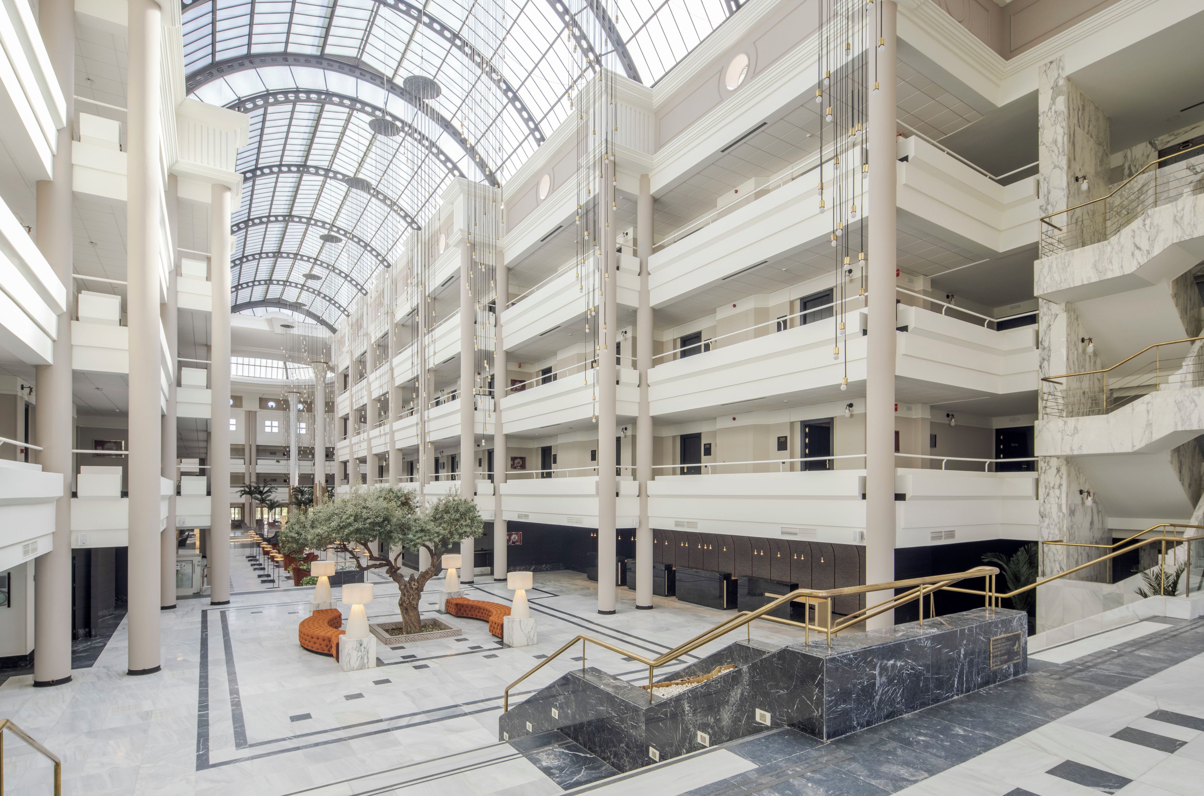 A multi-level atrium with a glass ceiling, marble flooring, and a central seating area with a tree.