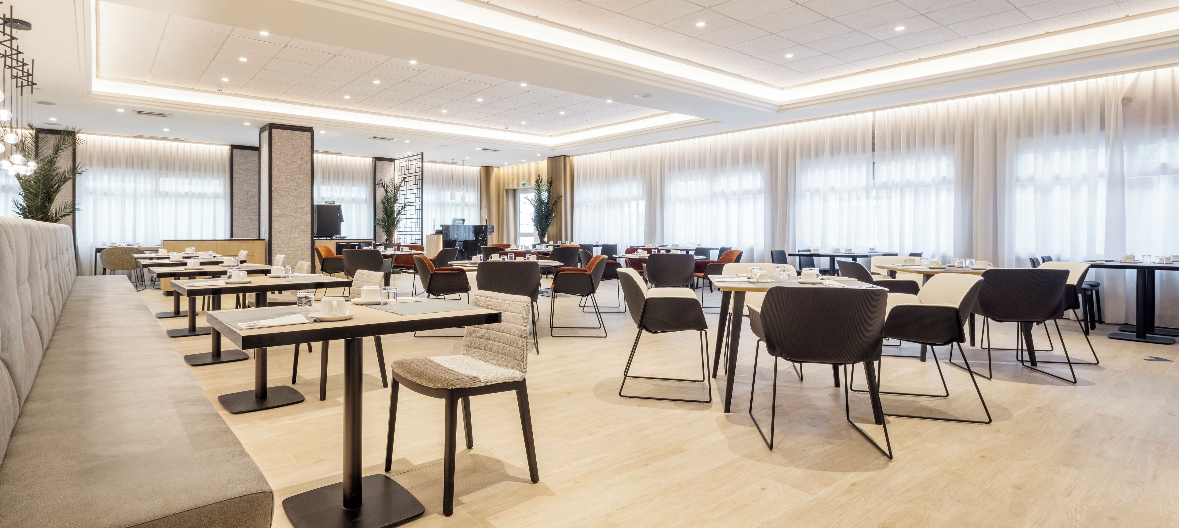 A spacious hotel dining area with multiple tables and chairs, large windows with curtains, and a modern ceiling design.