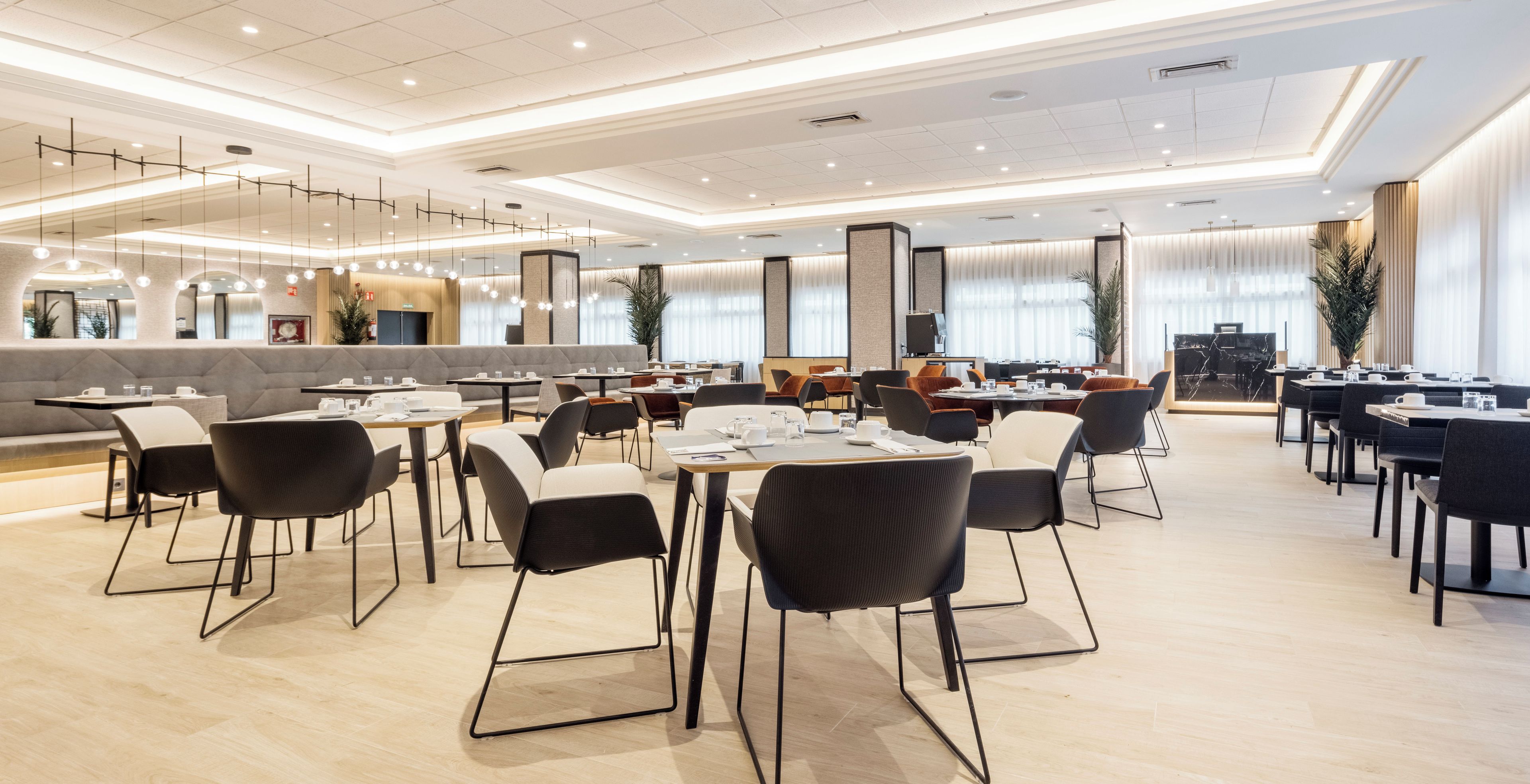 A modern dining area with multiple tables and chairs, a seating area with a sofa, and a large window providing natural light.
