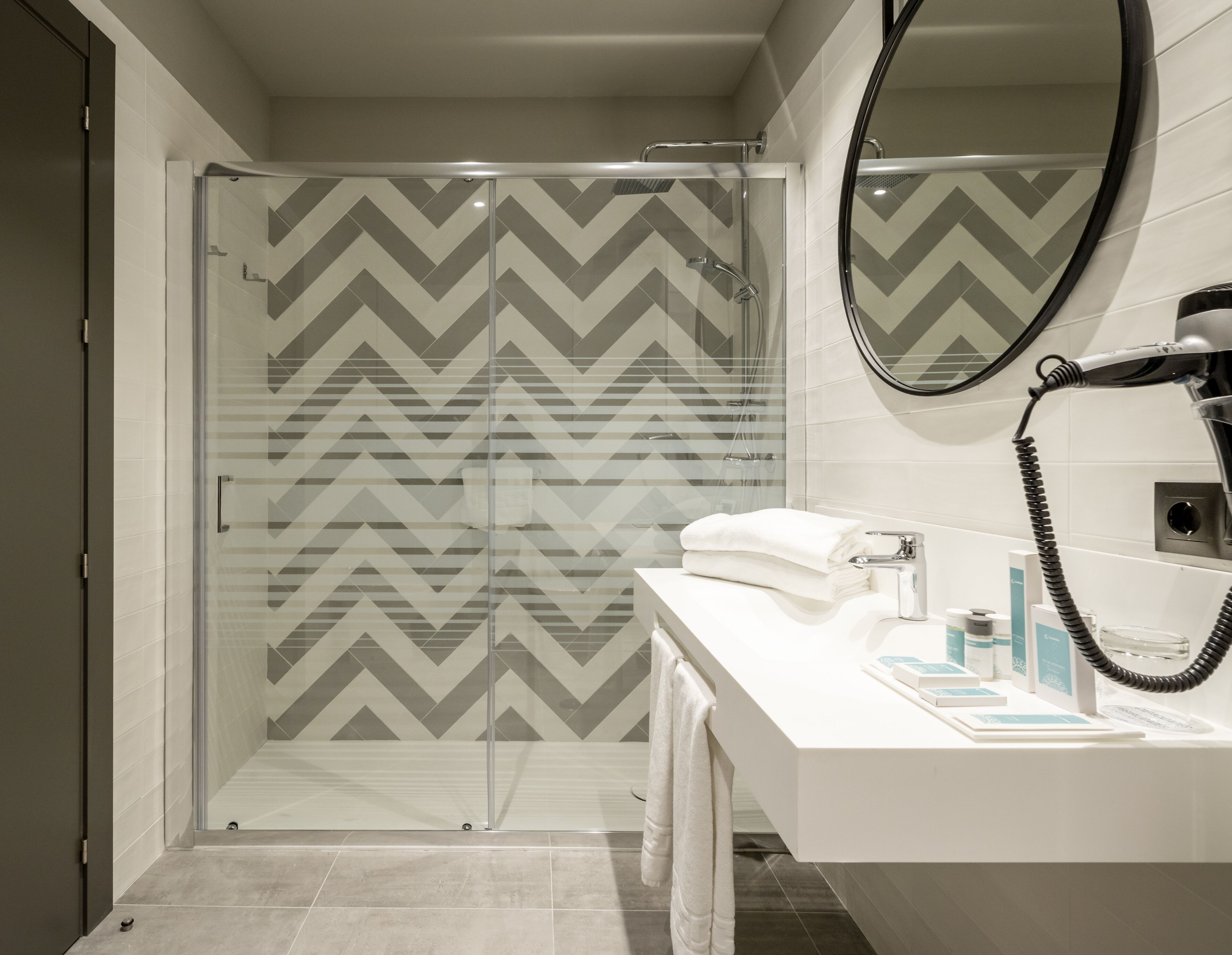 A modern bathroom with a glass shower enclosure, a white sink with toiletries, and a large mirror.