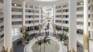 A spacious atrium with a central open area surrounded by multiple floors, featuring a glass dome ceiling and palm trees. - Hotel ILUNION Alcora Sevilla (San Juan de Aznalfarache)