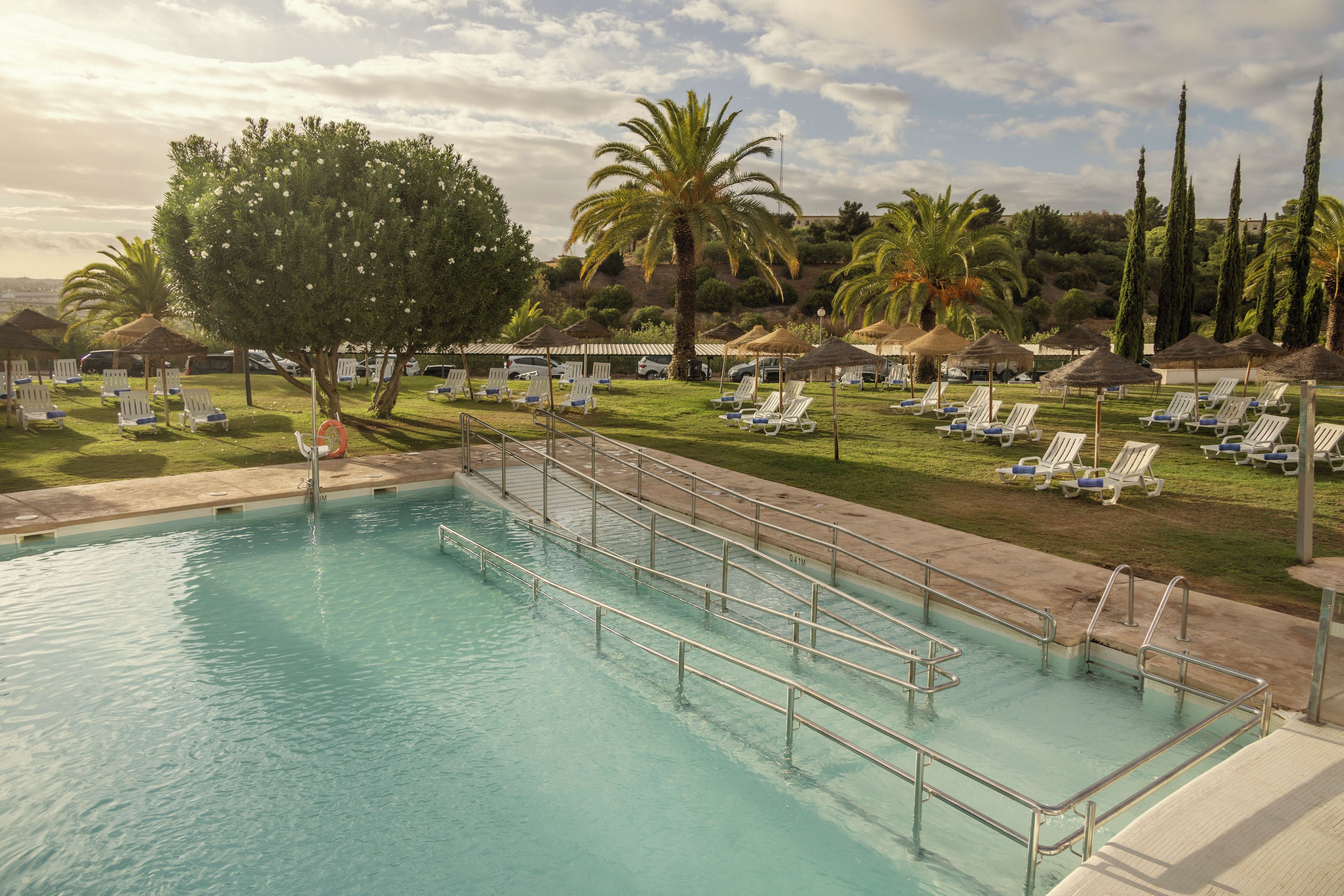 A swimming pool with sun loungers and umbrellas, surrounded by palm trees and a hilly landscape.