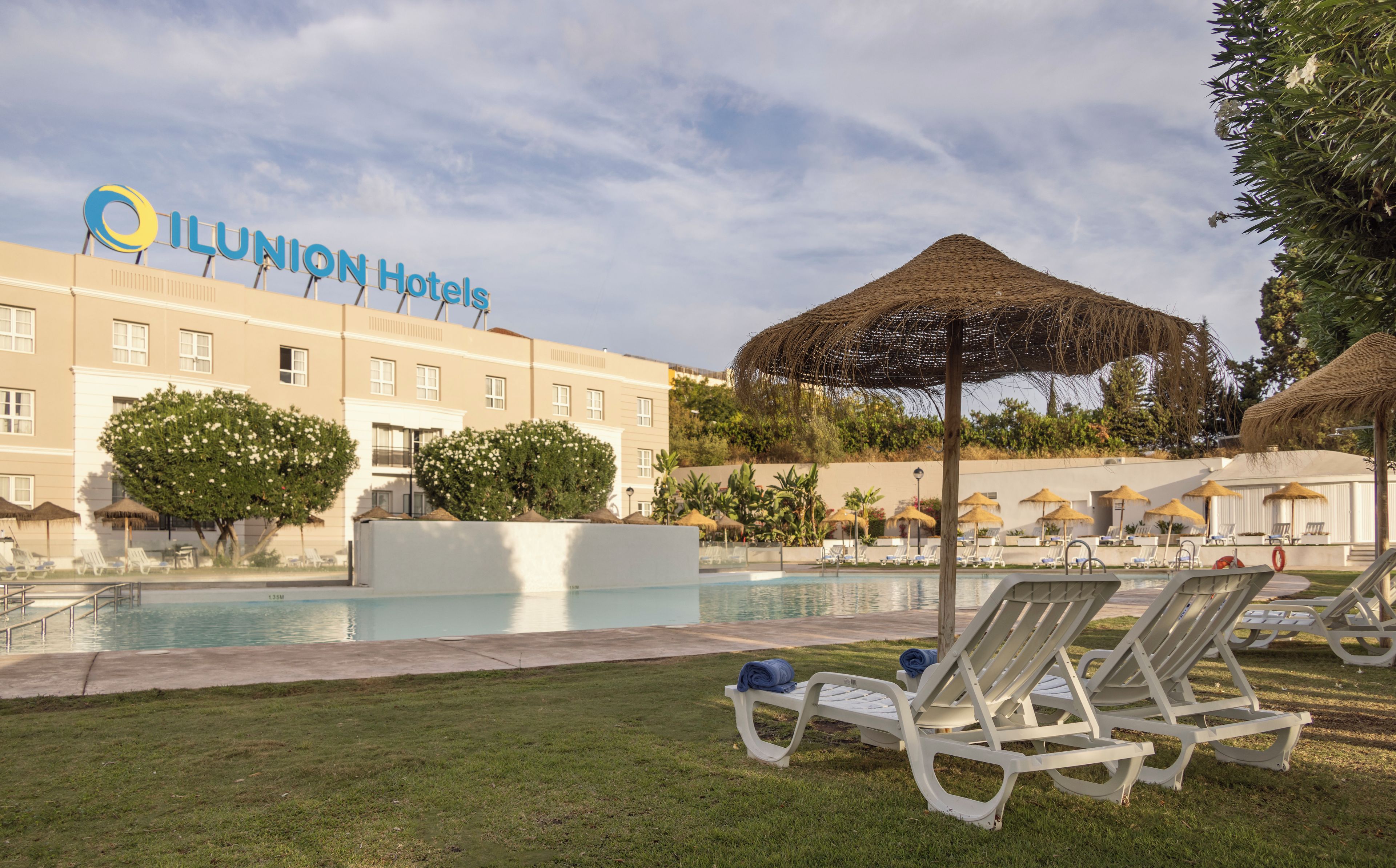 Un espace piscine d’hôtel avec des chaises longues, un parasol en chaume et une vue dégagée sur le bâtiment de l’hôtel en arrière-plan.
