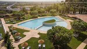 A large swimming pool surrounded by sun loungers and trees, with a view of a cityscape in the background. - Hotel ILUNION Alcora Sevilla (San Juan de Aznalfarache)