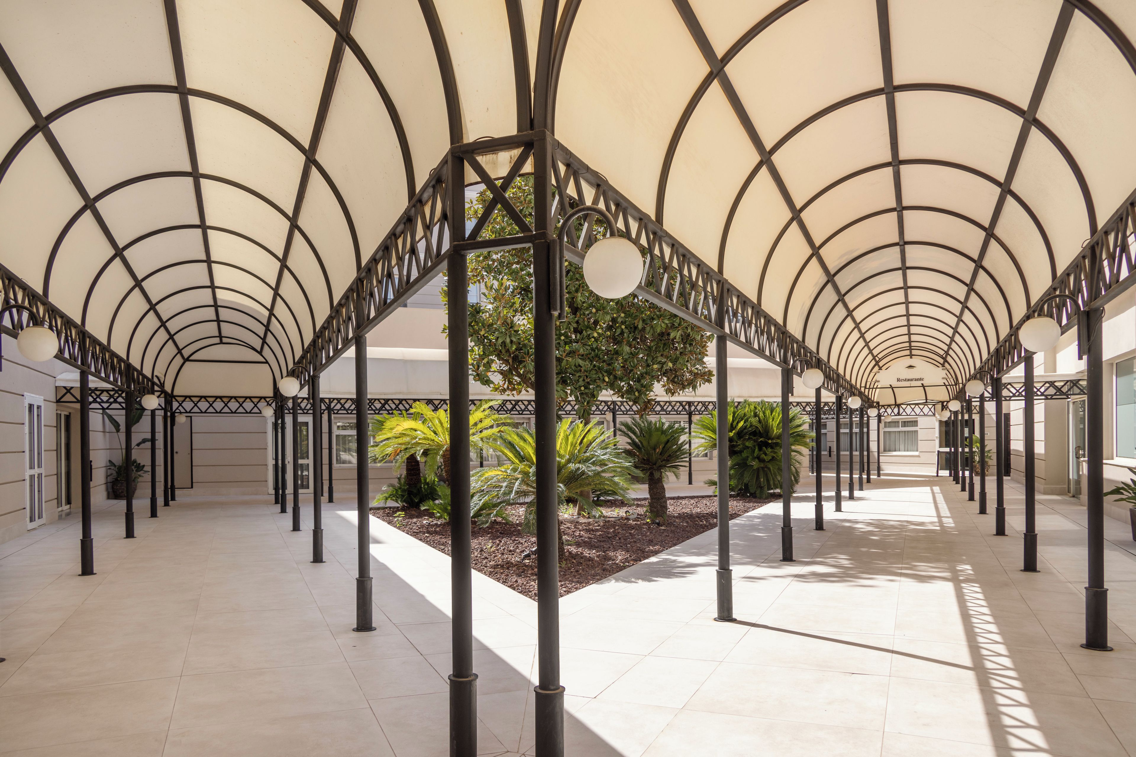 A covered walkway with a geometric metal framework, potted plants, and a tiled floor.