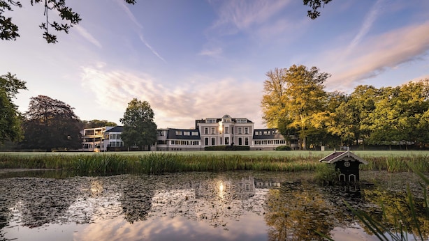 A large, multi-story building with a symmetrical facade, a small structure with a pointed roof to the right, and a body of water reflecting the building and trees. - Hotel Landgoed Lauswolt (Beetsterzwaag)