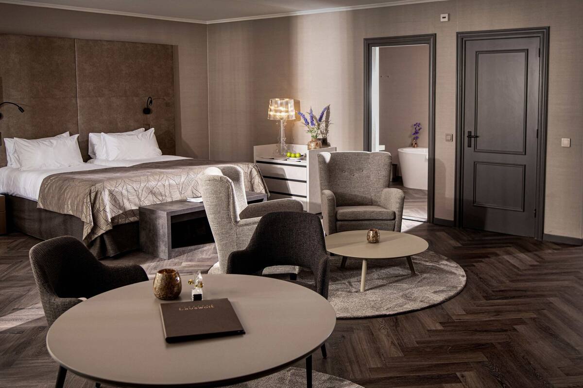 A modern hotel room with a large bed, two armchairs, a small table, and a bedside table with a lamp and flowers.