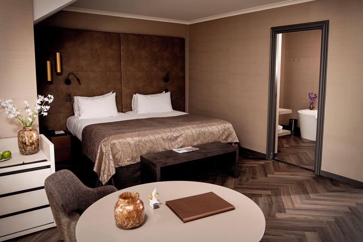 A hotel room with a large bed, a bedside table, a chair, a round table, and a mirror.