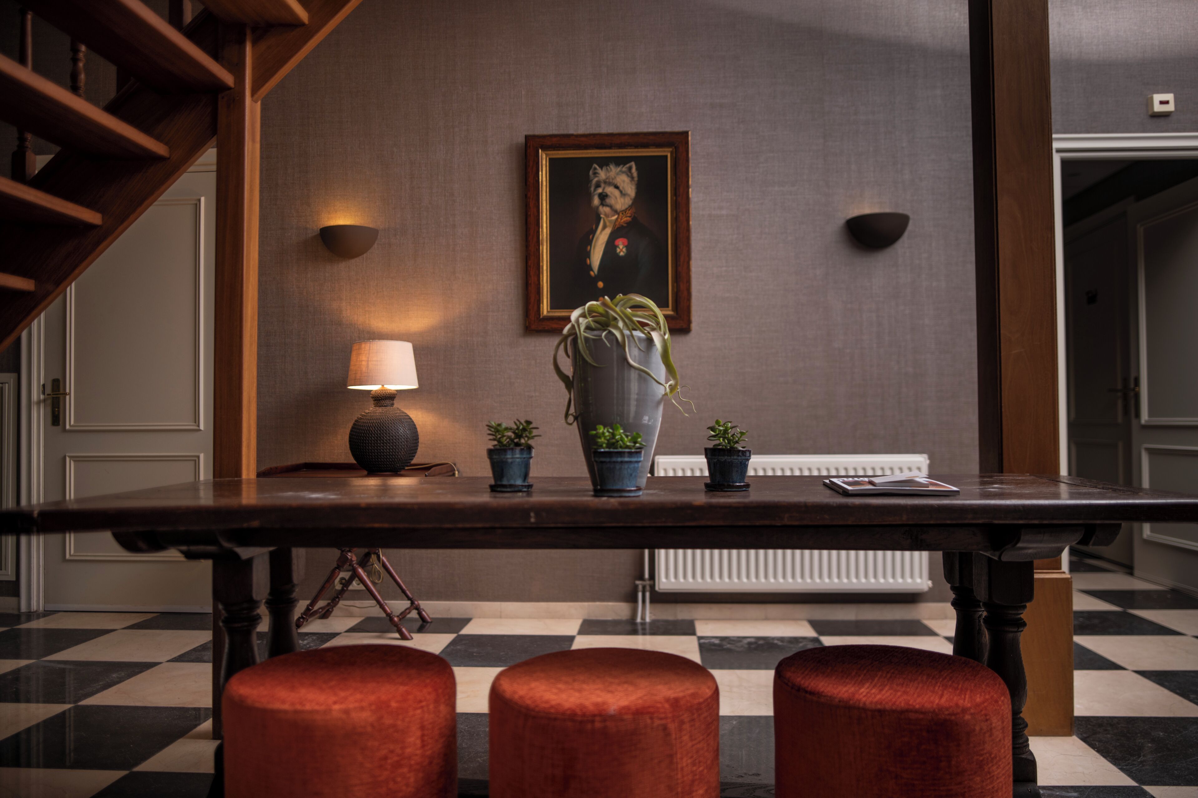 A checkered floor, a wooden table with potted plants, a lamp, and a framed portrait on the wall.