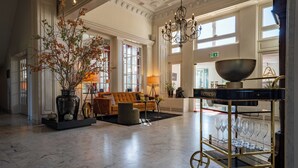 A spacious lobby with a marble floor, a sofa, a vase with branches, a chandelier, and a cart with wine glasses. - Hotel Landgoed Lauswolt (Beetsterzwaag)