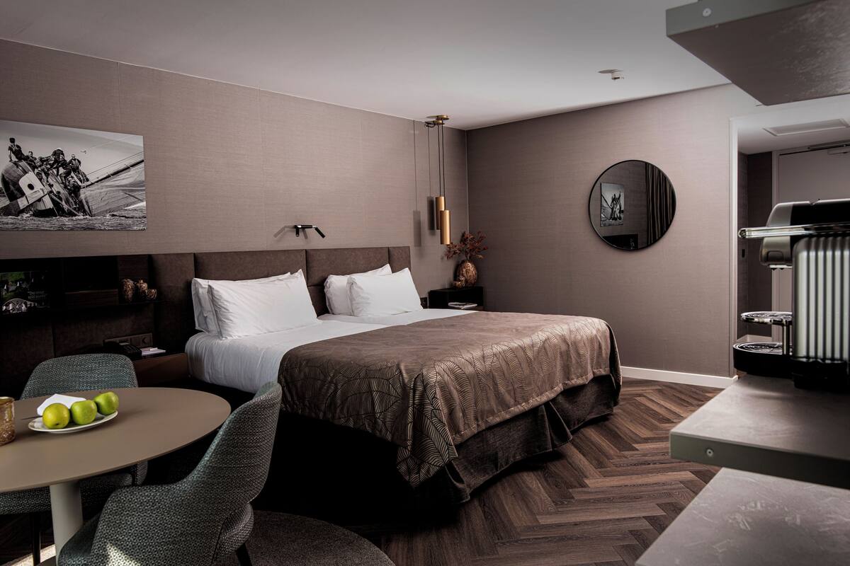 A modern hotel room with a large bed, a round table with apples, a chair, a small table with a vase, and a wall-mounted TV.