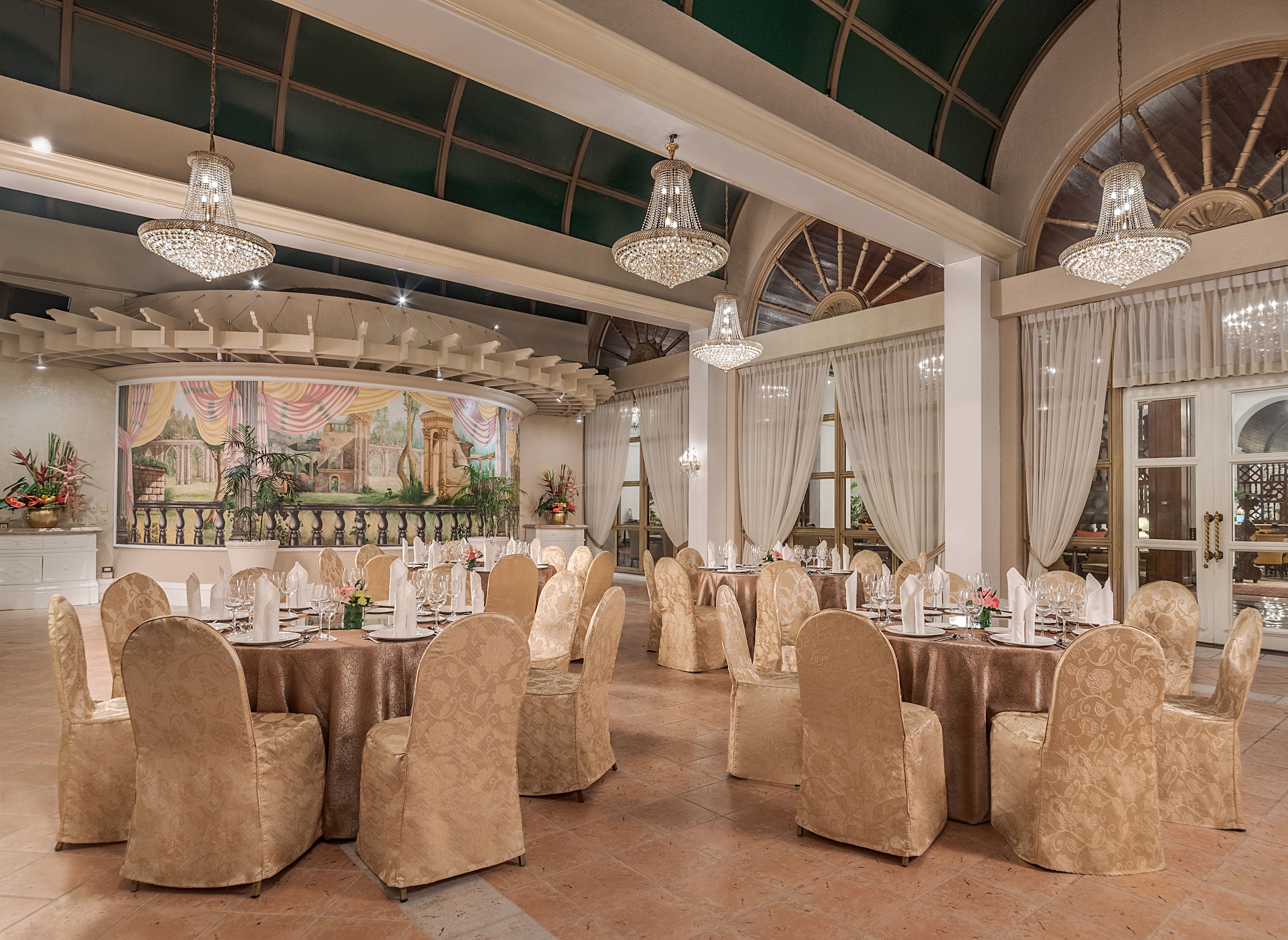 A banquet hall with round tables set for a formal event, featuring chandeliers and a mural on the wall.