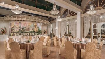 Un salón de banquetes con mesas redondas dispuestas para un evento formal, con candelabros y un mural en la pared.