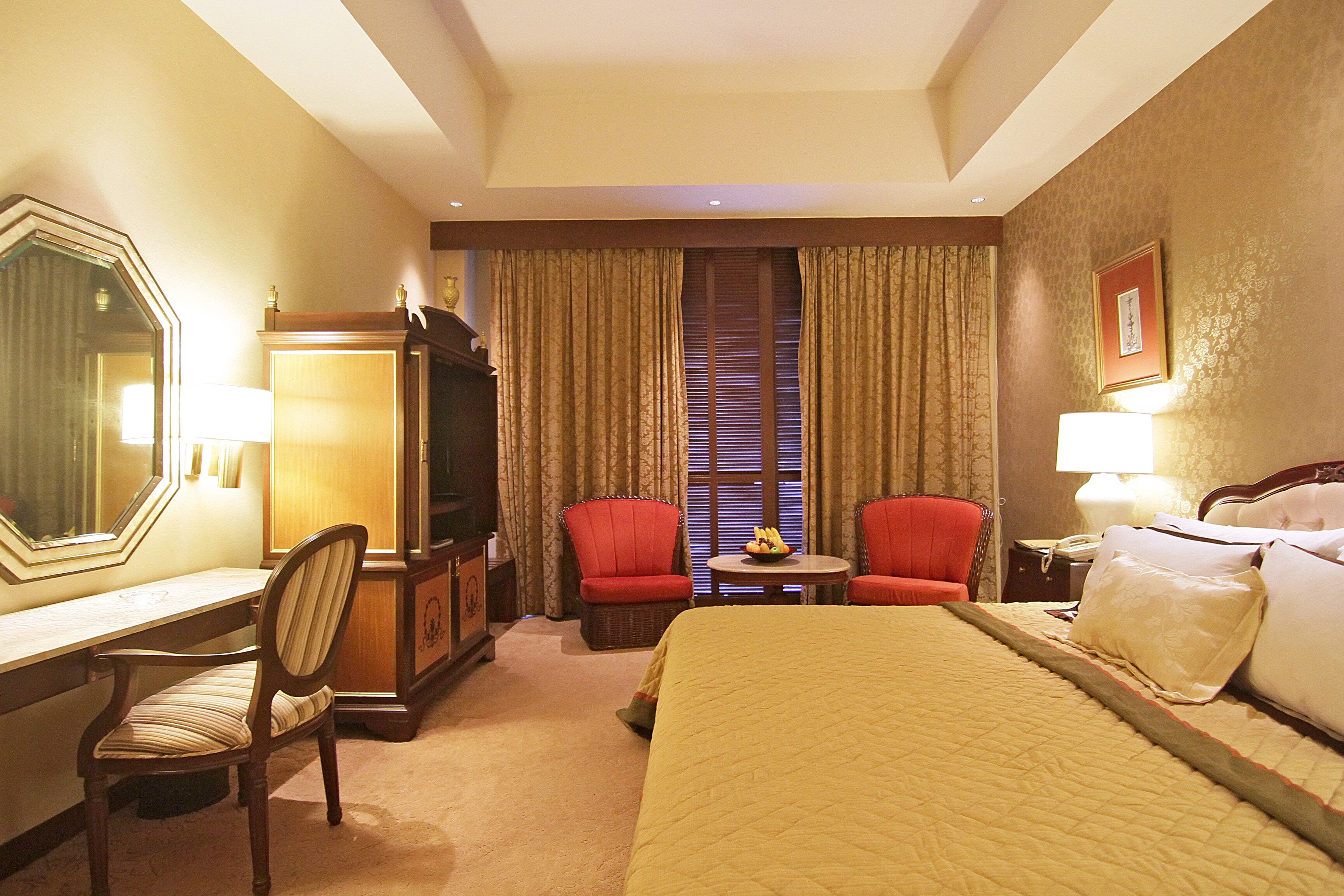 A hotel room with a bed, two red chairs, a desk, a dresser, and a small table with fruit.