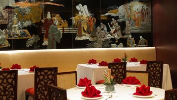 A restaurant with round tables set with white tablecloths, red napkins, and traditional Chinese artwork on the walls.