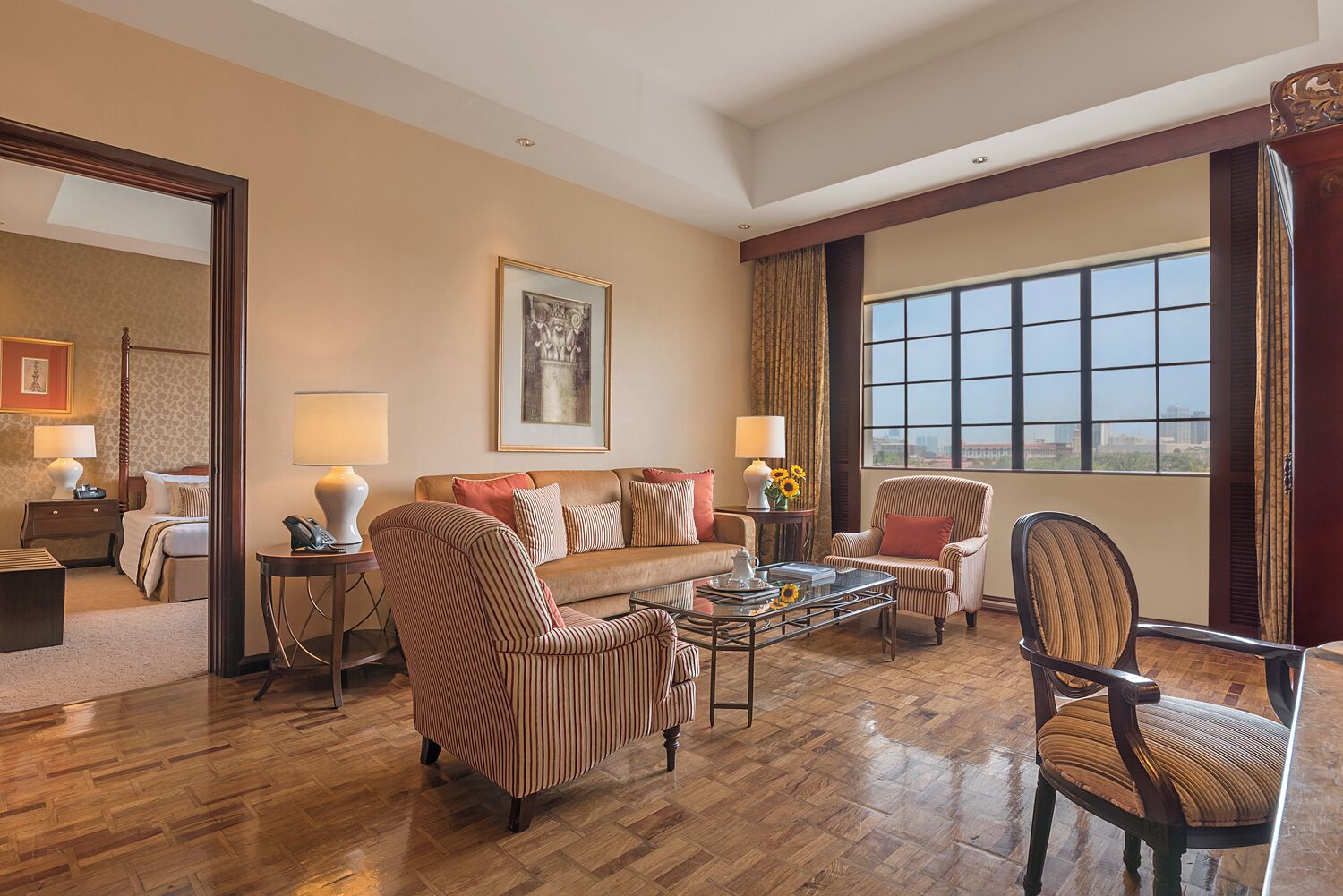 A spacious living room with a large window, a four-poster bed, and a comfortable seating area with striped armchairs and a sofa.