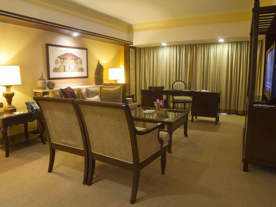 A hotel room with a sofa, two armchairs, a glass coffee table, a desk with a chair, a television, and a painting on the wall.