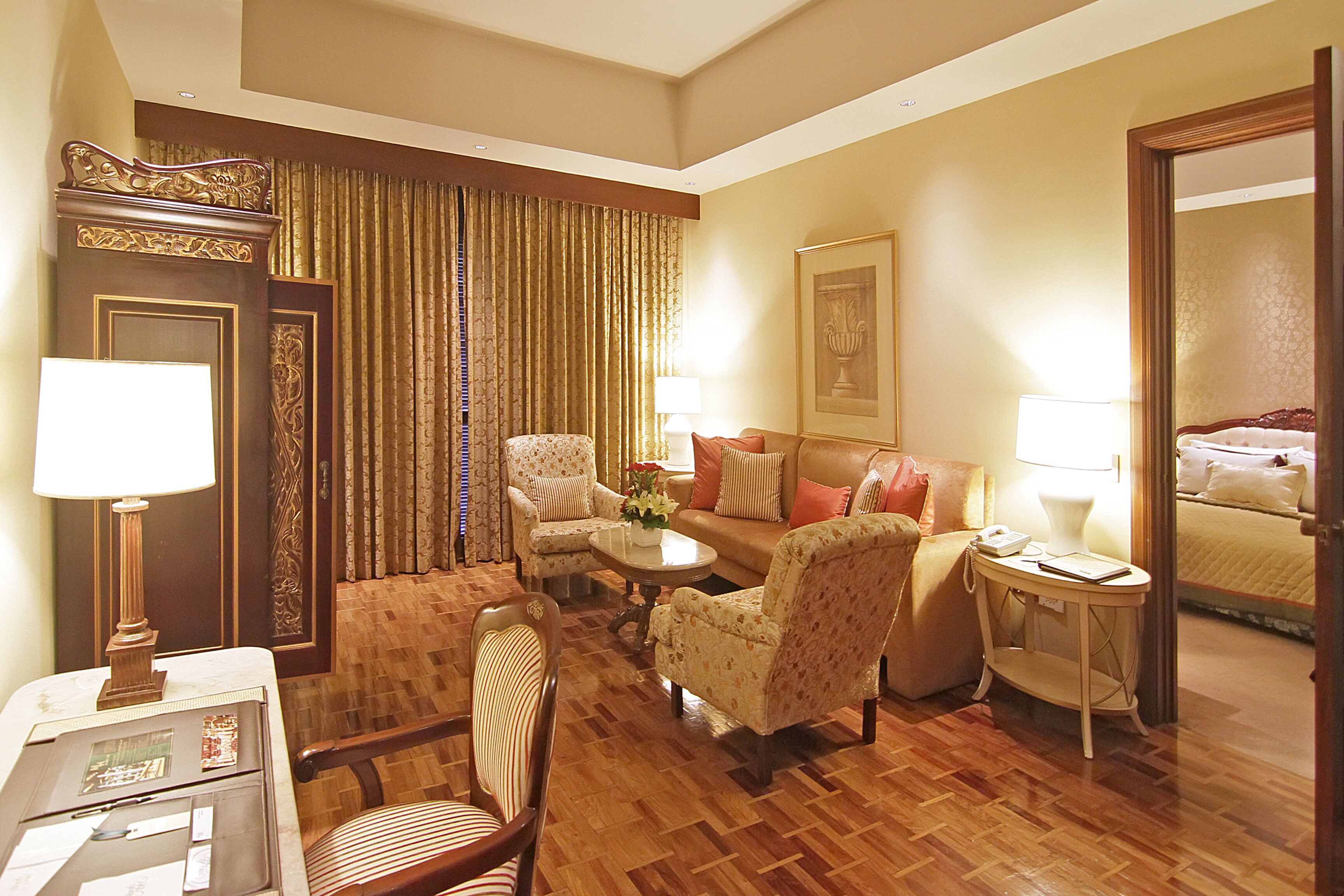 A hotel room with a wooden floor, a sofa, two armchairs, a coffee table, a side table with a lamp, a desk with a lamp, a door with curtains, and a bed with a headboard.