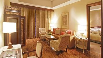 A hotel room with a wooden floor, a sofa, two armchairs, a coffee table, a side table with a lamp, a desk with a lamp, a door with curtains, and a bed with a headboard.