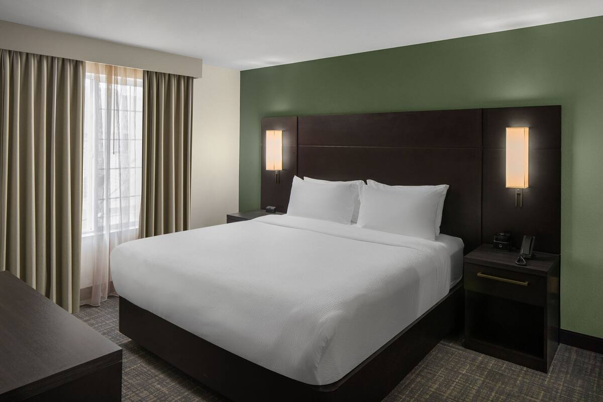 A hotel room with a large bed, two bedside tables with lamps, a nightstand, and a window with curtains.