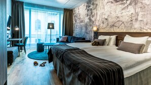 Ein modernes Hotelzimmer mit einem großen Bett, einem Schreibtisch, einer Sitzecke und Blick auf die Stadt durch große Fenster.