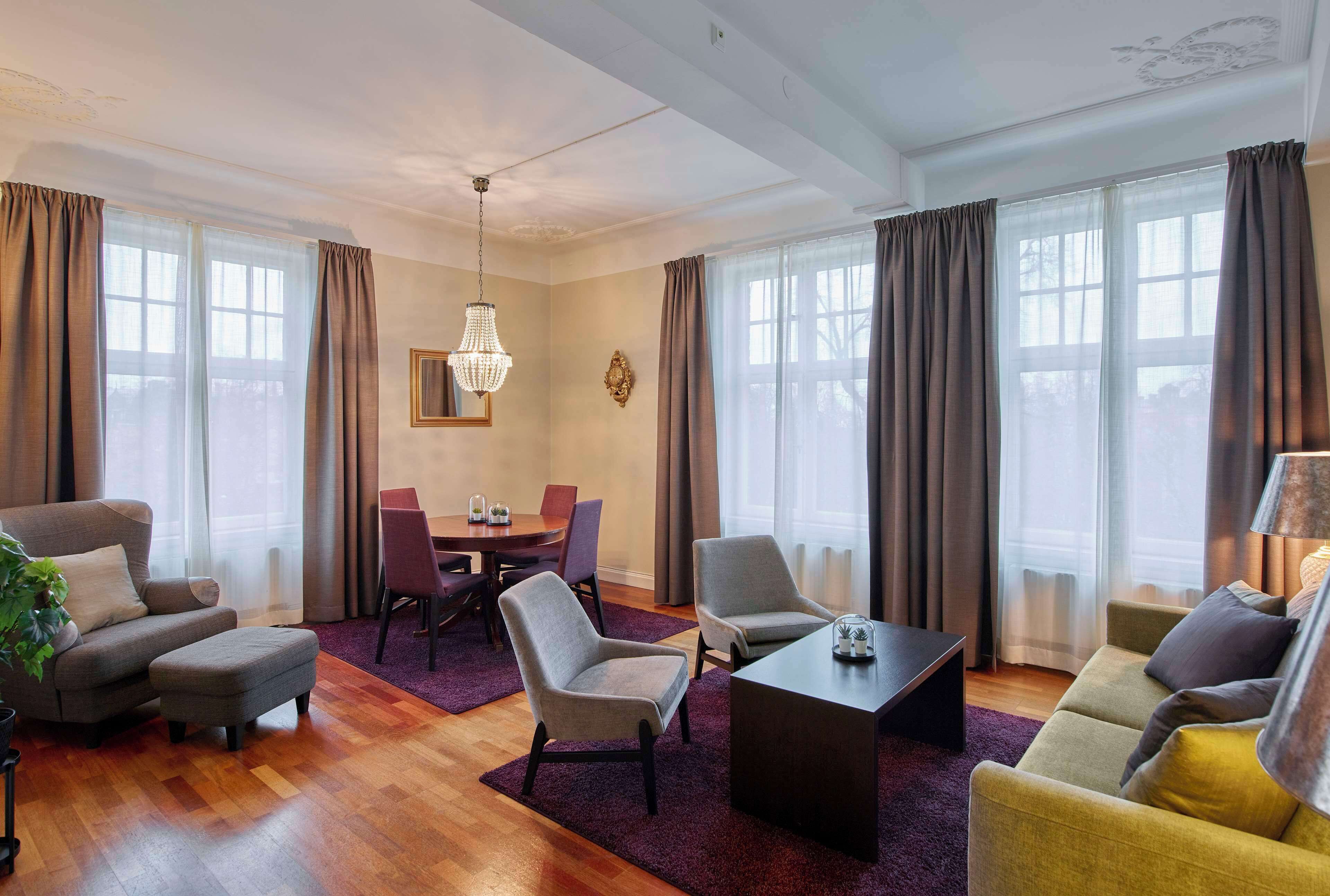 Una sala moderna con sofá, sillón, reposapiés y una mesa de centro. Se observa una mesa de comedor con sillas y una araña.