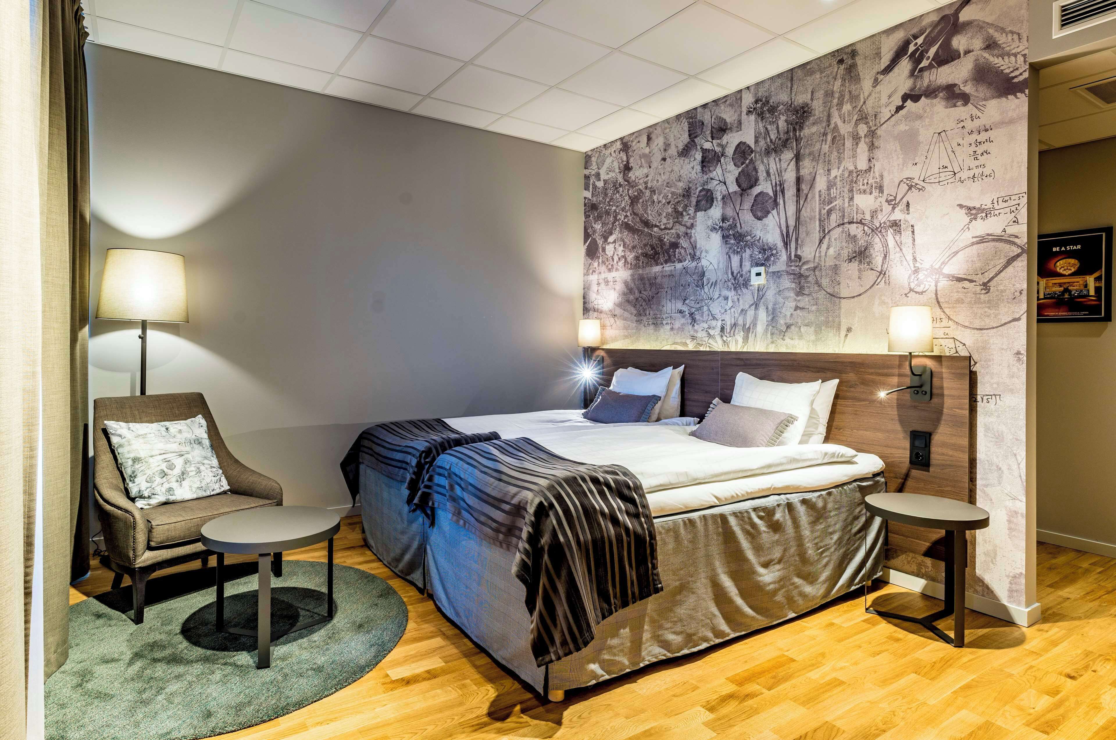 A modern hotel room with a large bed, a chair, a small table, and a wall mural.