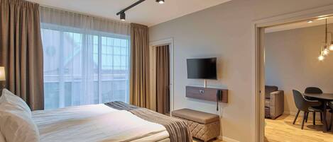 A modern hotel room with a large bed, a flat-screen TV, a desk with a chair, and a dining area.