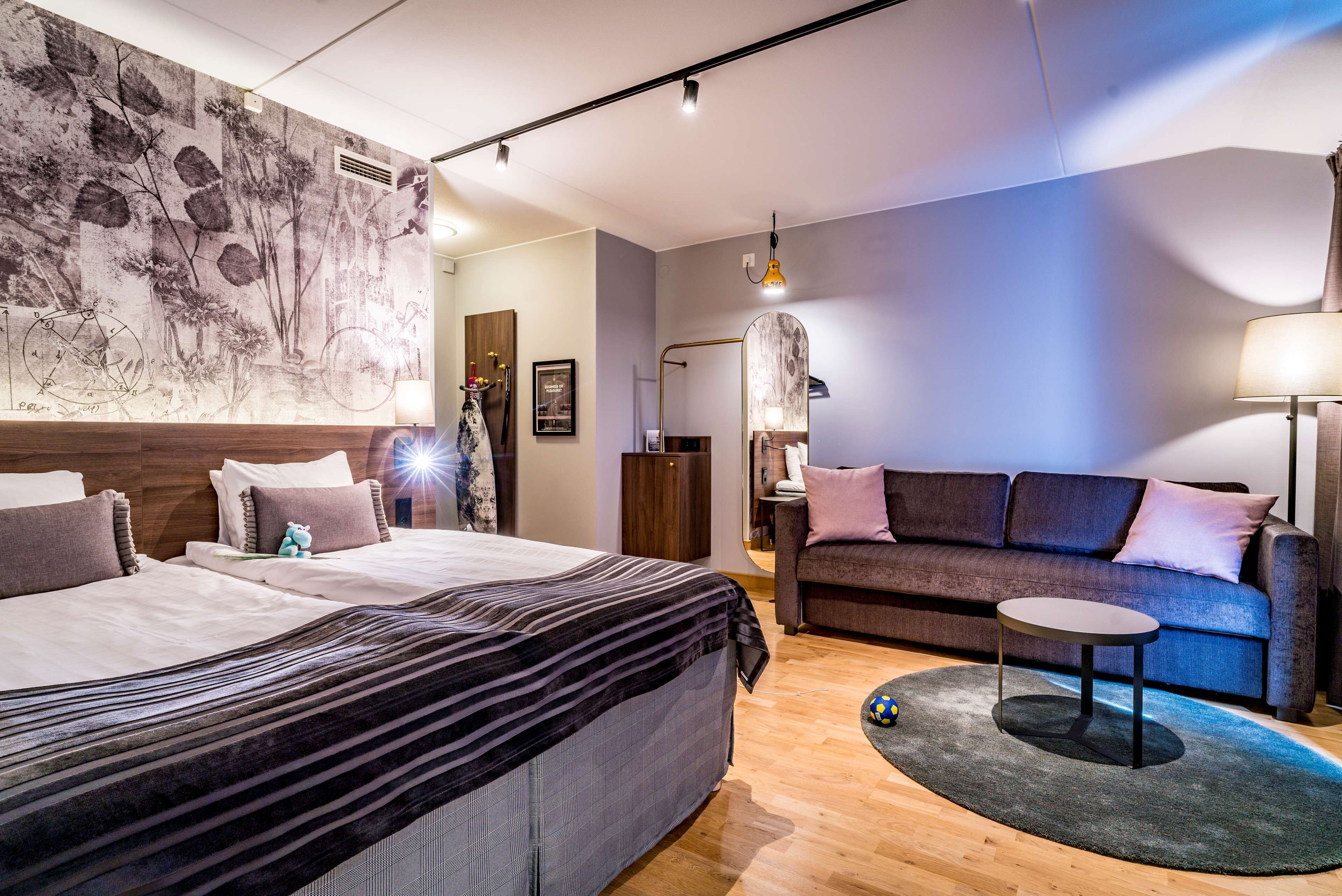 A modern hotel room with a large bed, a sofa, a small table, and a mirror.