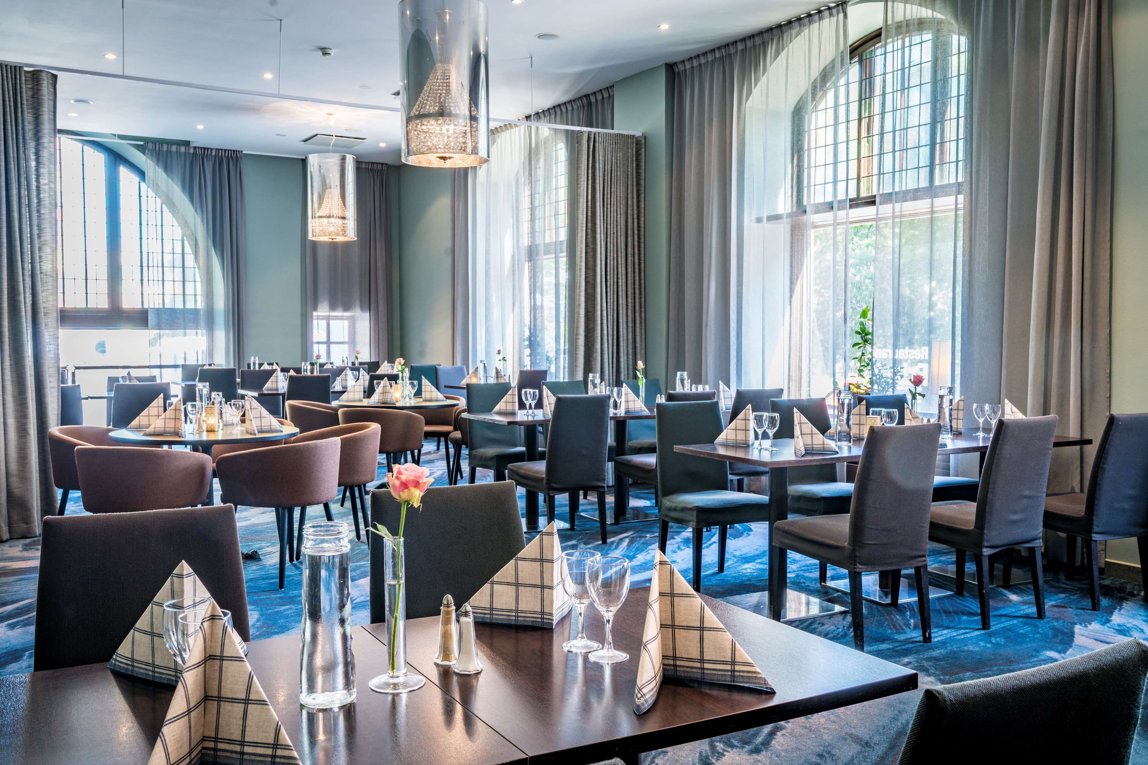 Un'ampia sala da pranzo ben illuminata, con tavoli apparecchiati, grandi finestre con tende e un lampadario.