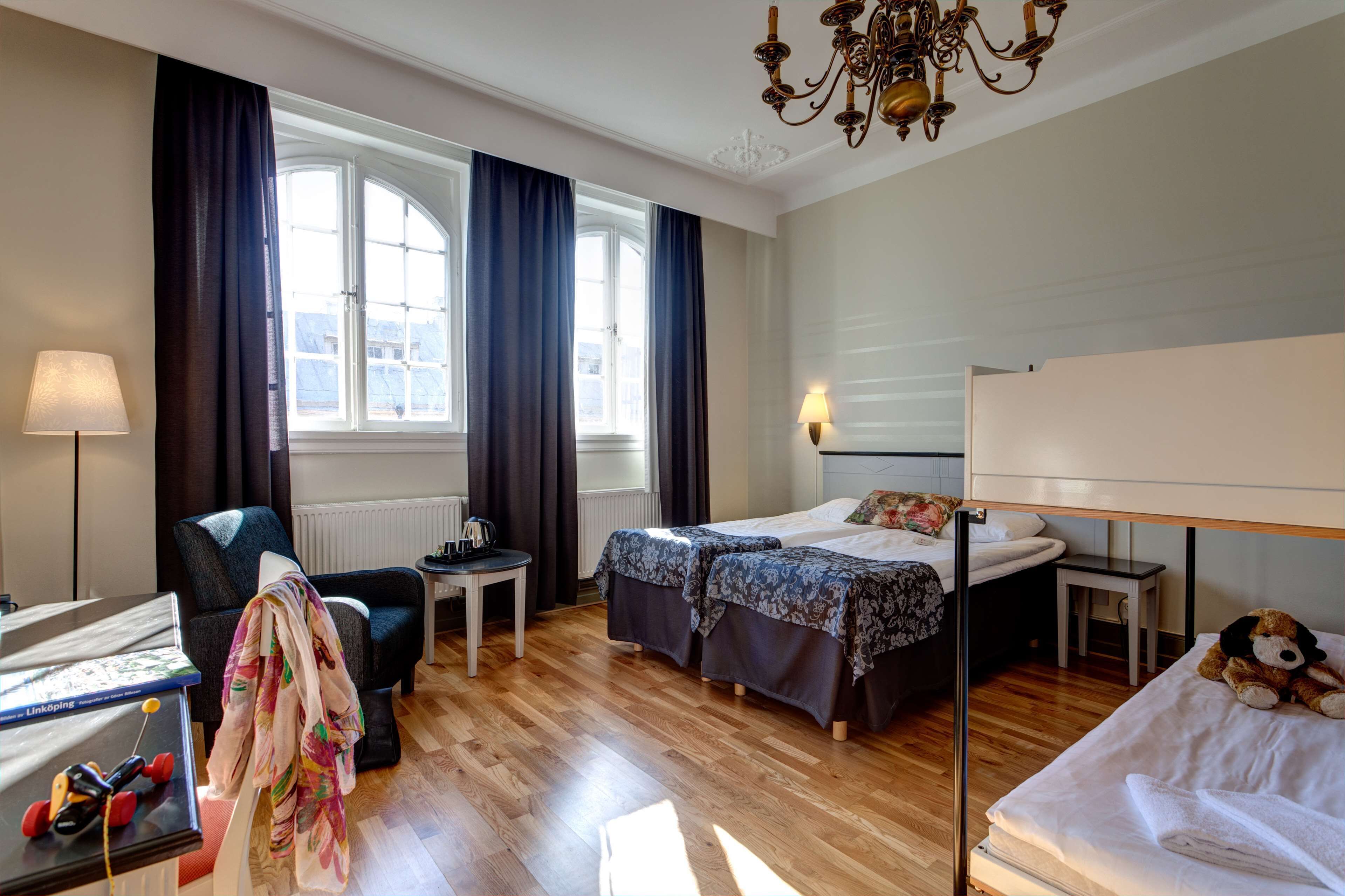 Habitación de hotel con cama, dos mesitas de noche, una silla, un escritorio, una lámpara, un candelabro y una ventana con cortinas.