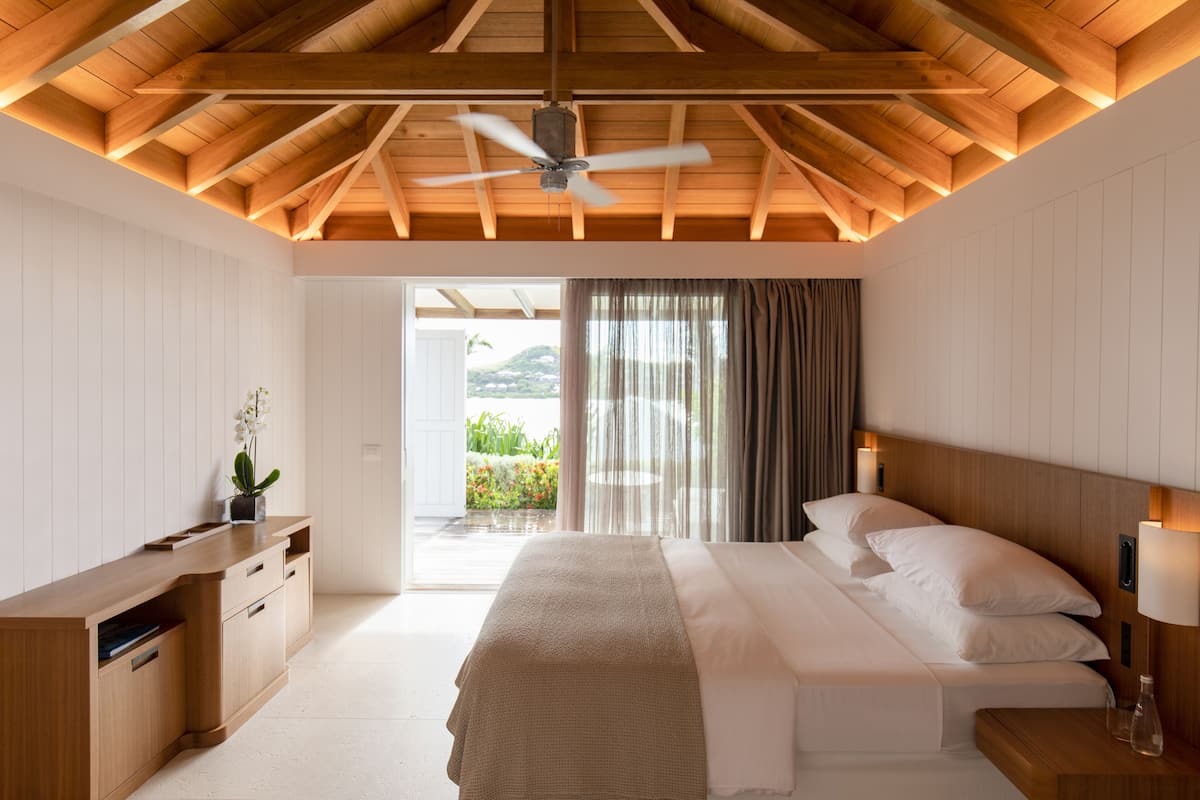 A bedroom with a wooden ceiling, a bed with white linens, a nightstand, a desk with drawers, and a view of greenery outside.