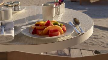 A table with a plate of fruit and a cup, set on a beach with a view of the ocean.