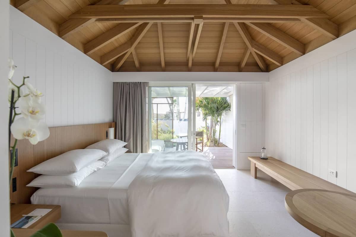 A bedroom with a large bed, a wooden bench, and a view of the outdoors.