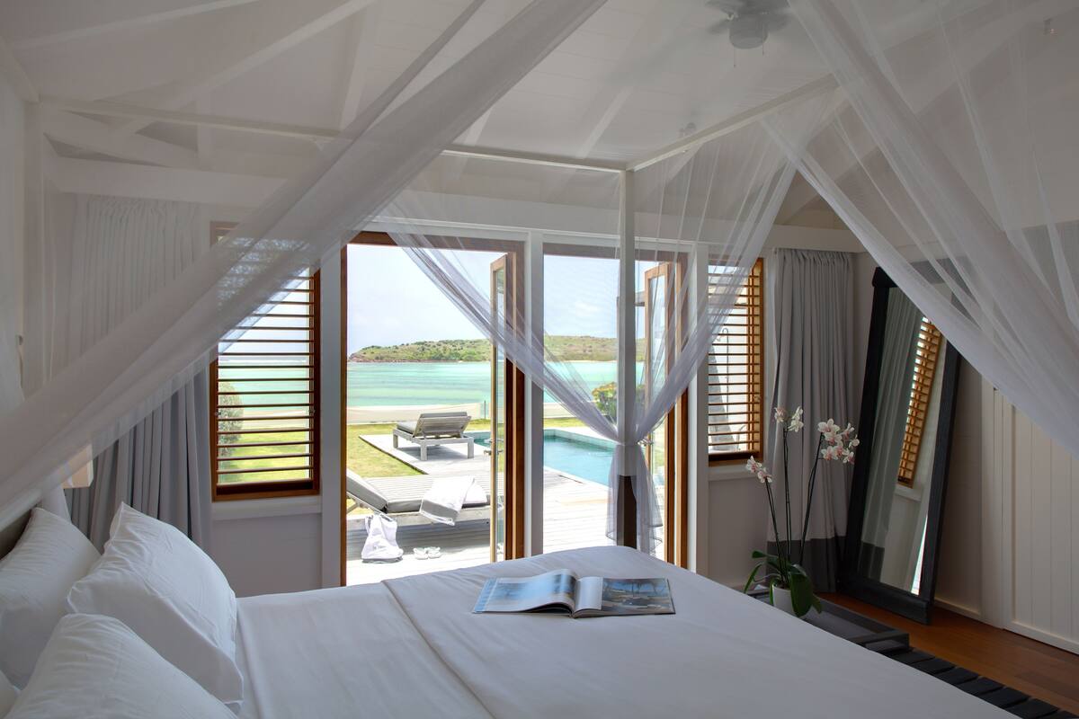 A bedroom with a large bed, a view of a pool and lounge chairs, and sheer curtains.
