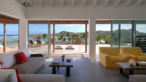 A spacious living area with a large window overlooking a beach and ocean, furnished with a sofa, armchairs, and a coffee table. - Le Sereno (St. Barthelemy)