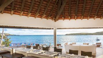 Un área de comedor al aire libre con mesas y sillas, con vista a un cuerpo de agua y una isla distante.