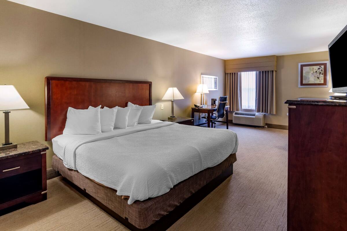 A hotel room with a large bed, two bedside tables with lamps, a desk with a chair, a flat-screen TV, and a window with curtains.