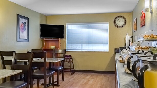 A dining area with wooden chairs and tables, a television mounted on the wall, a window with blinds, and a breakfast bar with a coffee pot. - Quality Inn & Suites Vancouver North (Vancouver)