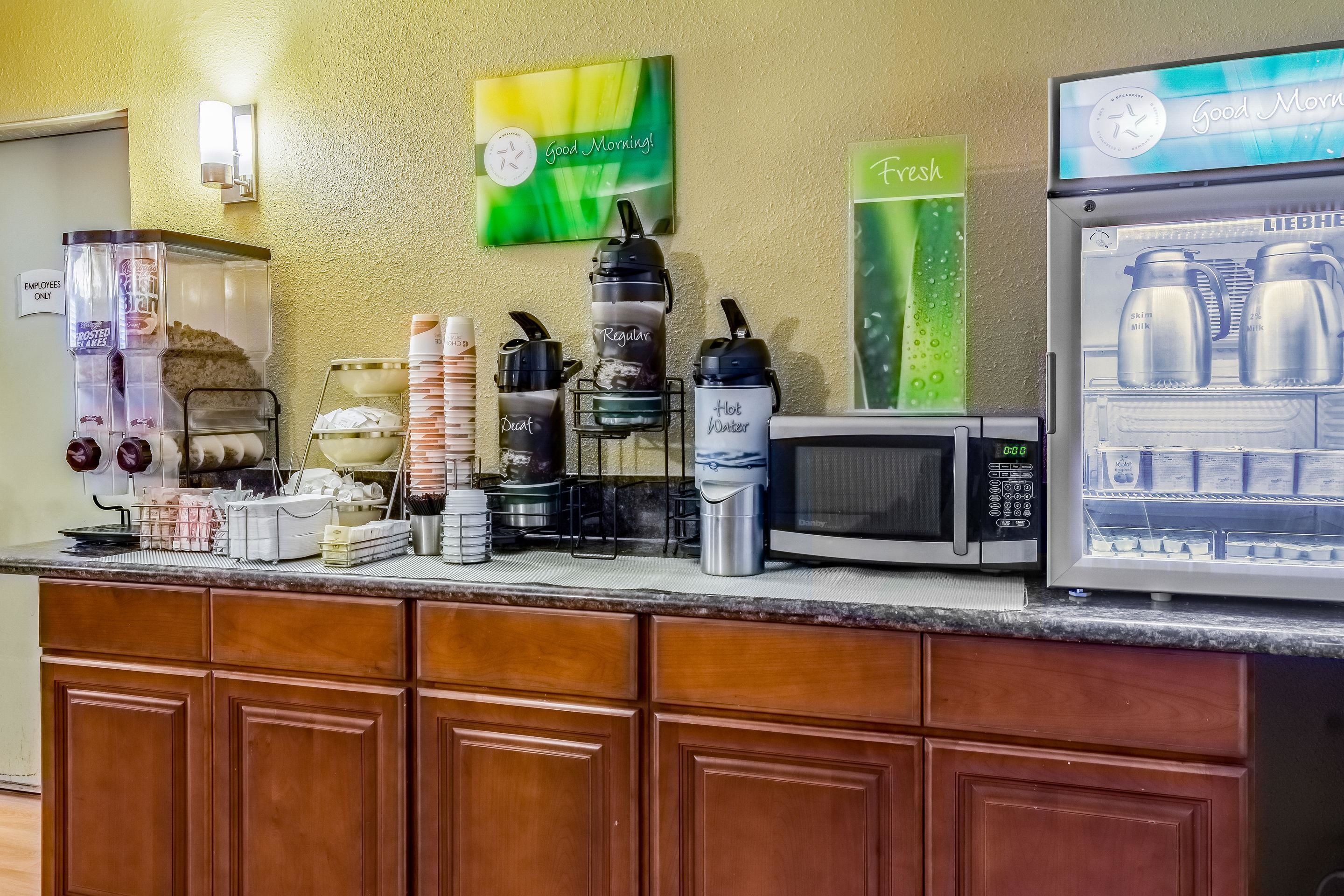 A hotel breakfast area with a coffee station, a microwave, and a refrigerator displaying milk cartons.