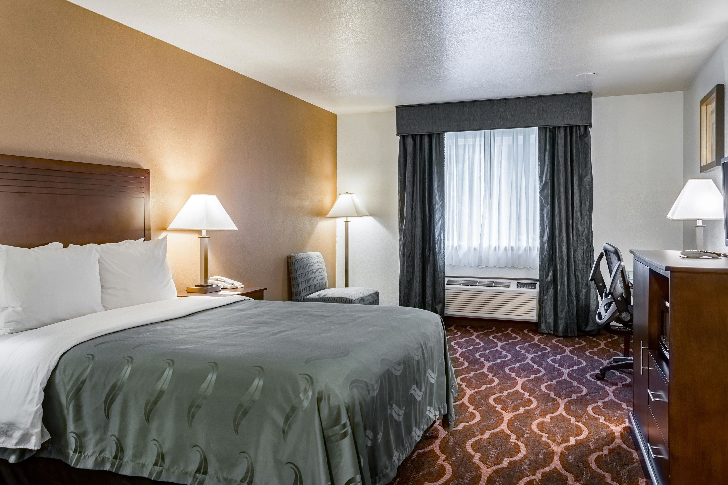 A hotel room with a large bed, two bedside lamps, a desk with a chair, an air conditioning unit, and a window with curtains.